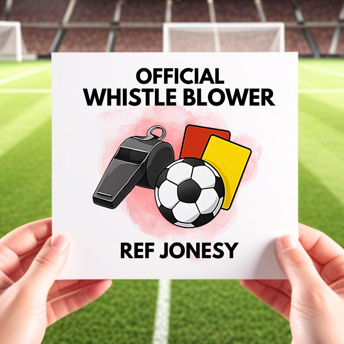Close-up of a hand holding a card with the text "Official Whistle Blower" and an illustration of a whistle, soccer ball, red card, and yellow card, set against a blurred soccer field background.
