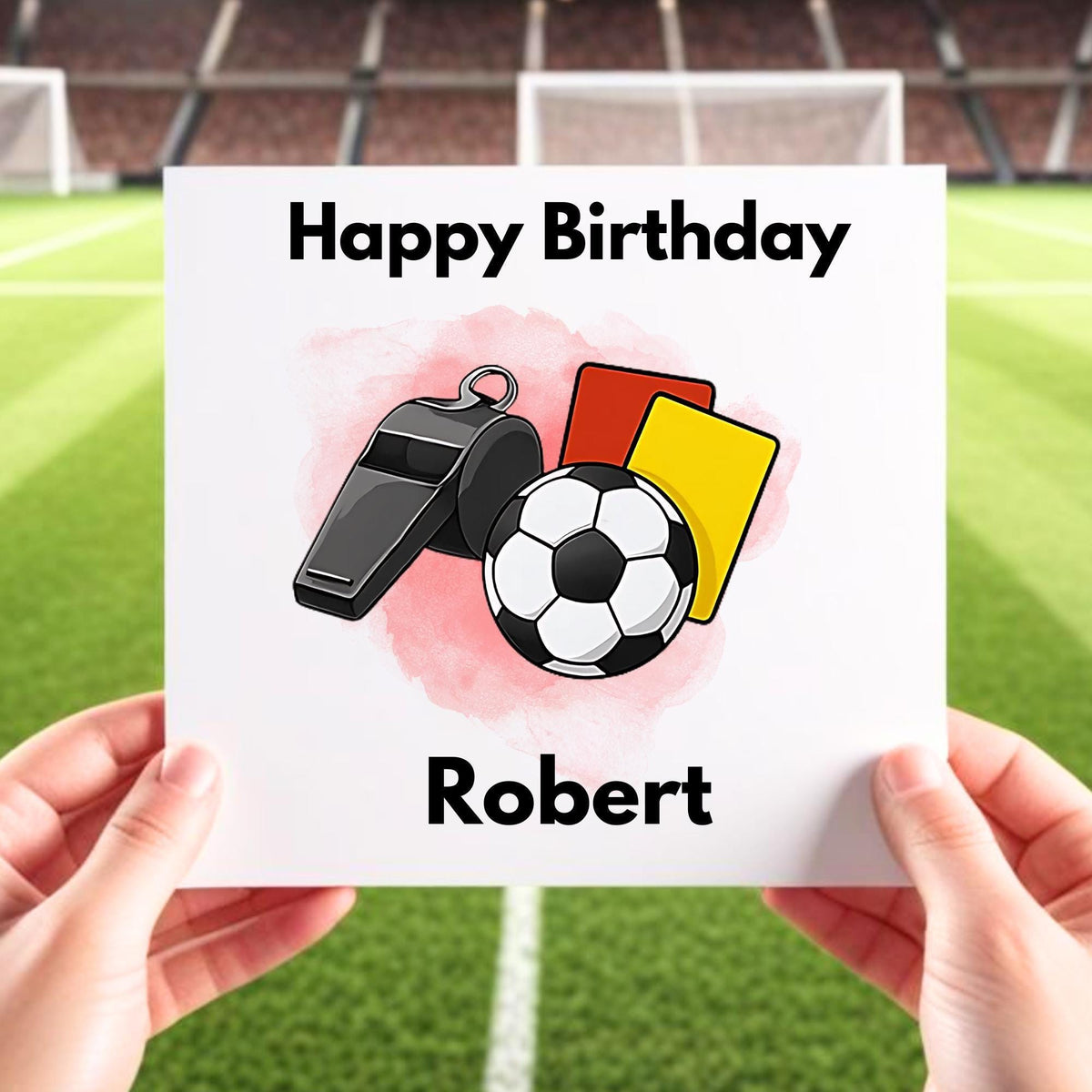 A close-up shot of a pair of hands holding a personalized "Happy Birthday" card with a referee design against the backdrop of a blurred soccer field.  The card features a soccer ball, whistle, and referee cards, and the name "Robert" is printed at the bottom.