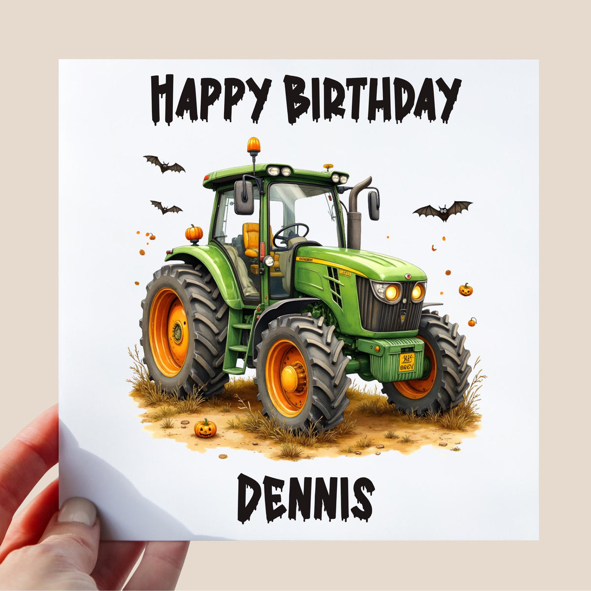 A hand holding a square birthday card featuring a green tractor decorated with pumpkins and bats, with the words "Happy Birthday" above and the name "Dennis" below.