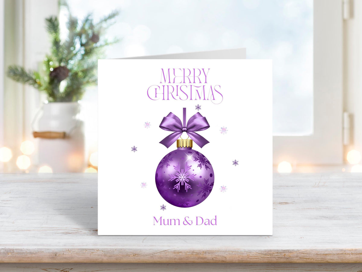 A square white Christmas card featuring a purple bauble with a bow at the top and snowflakes. The words "Merry Christmas" are above the bauble, and "Mum & Dad" is below. The card is displayed on a wooden surface, with a blurry, festive background including a plant and fairy lights.
