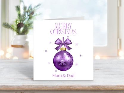 A square white Christmas card featuring a purple bauble with a bow at the top and snowflakes. The words "Merry Christmas" are above the bauble, and "Mum & Dad" is below. The card is displayed on a wooden surface, with a blurry, festive background including a plant and fairy lights.