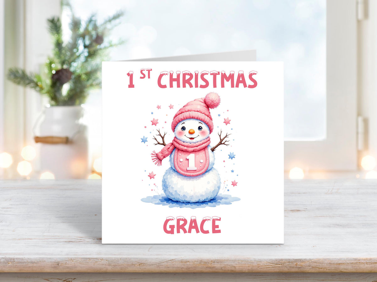 Personalised baby girl’s first Christmas card with a cute snowman wearing a pink hat and scarf, holding a “1” badge, with pink and blue stars and the name “Grace” at the bottom.