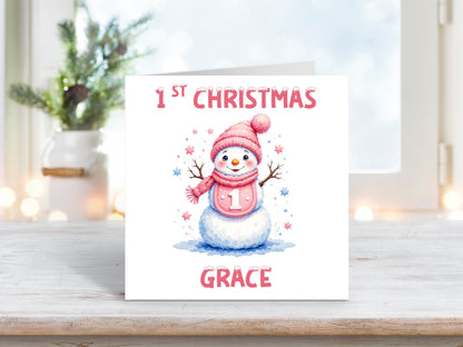 Personalised baby girl’s first Christmas card with a cute snowman wearing a pink hat and scarf, holding a “1” badge, with pink and blue stars and the name “Grace” at the bottom.