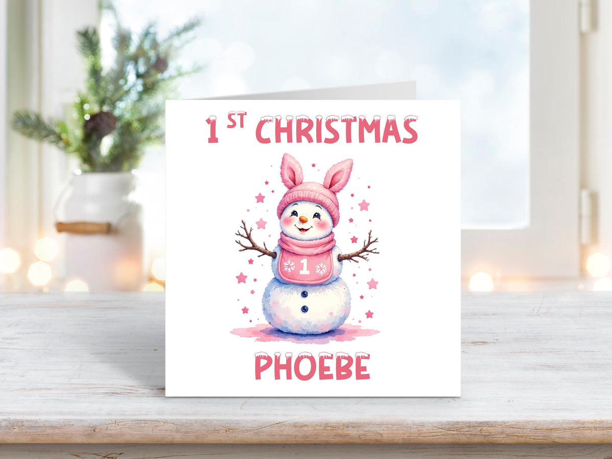 Personalised baby girl’s first Christmas card featuring a cute snowman with a pink bunny-eared hat and scarf, surrounded by pink stars, and the name “Phoebe” at the bottom.