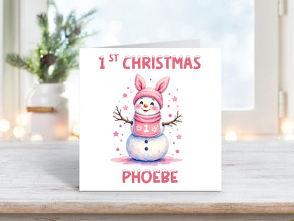 Personalised baby girl’s first Christmas card featuring a cute snowman with a pink bunny-eared hat and scarf, surrounded by pink stars, and the name “Phoebe” at the bottom.