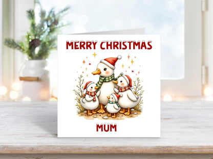 Christmas card featuring a family of ducks in Santa hats and scarves, surrounded by festive tree branches and sparkles, with “Merry Christmas Mum” in red text.