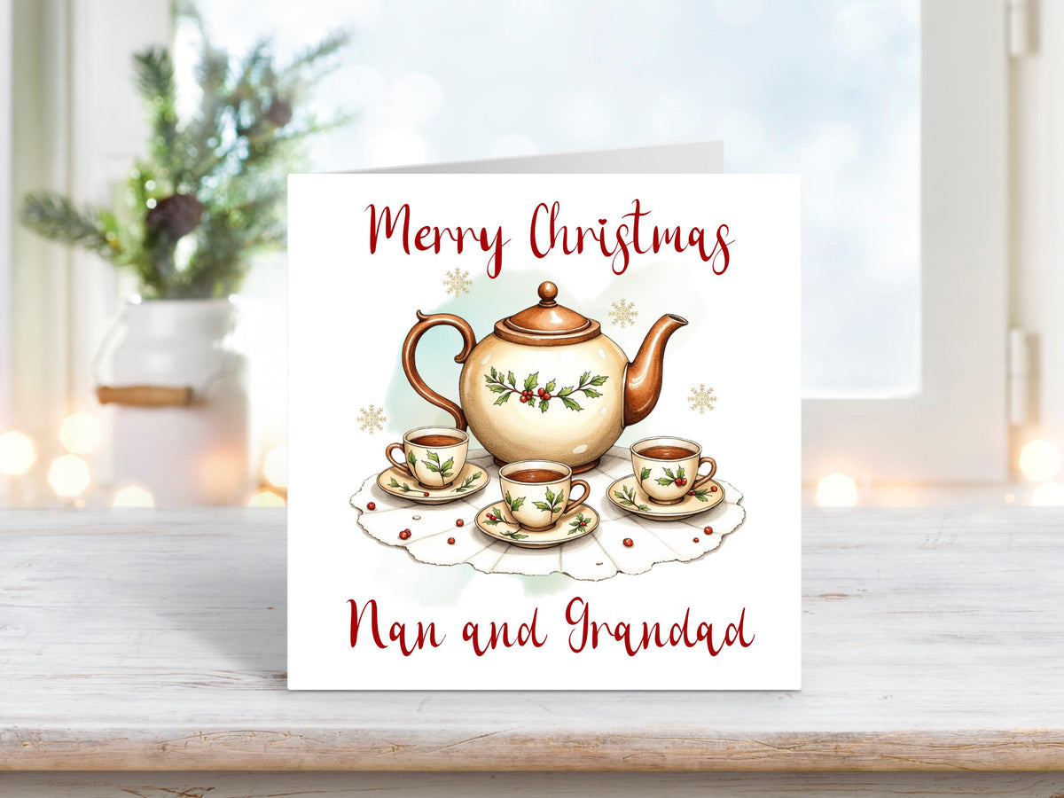 Christmas card featuring a festive teapot and matching teacups decorated with holly and berries on a lace doily, with the words “Merry Christmas Nan and Grandad” on a white background.