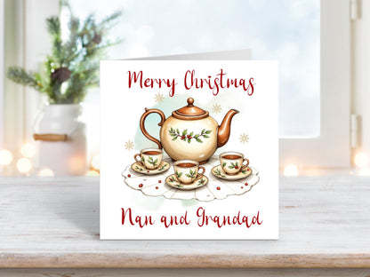 Christmas card featuring a festive teapot and matching teacups decorated with holly and berries on a lace doily, with the words “Merry Christmas Nan and Grandad” on a white background.