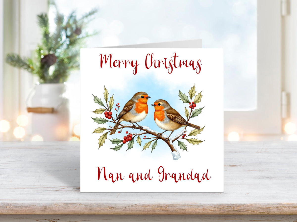 Christmas card featuring two robins perched on a holly branch with red berries and snow, under the text “Merry Christmas Nan and Grandad,” on a clean white background.