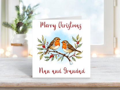 Christmas card featuring two robins perched on a holly branch with red berries and snow, under the text “Merry Christmas Nan and Grandad,” on a clean white background.