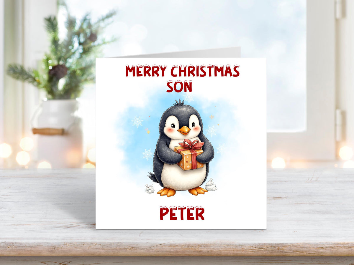 Christmas card featuring a smiling penguin holding a wrapped present against a soft blue snowy background, with the words “Merry Christmas Son” and personalised name “Peter” on a white background.