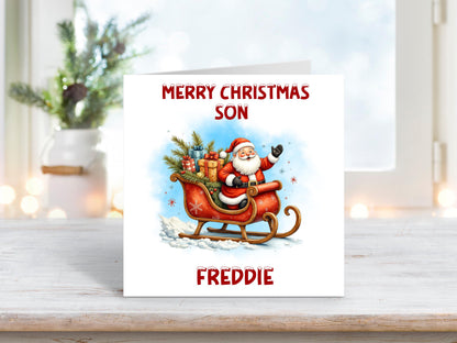 Christmas card featuring Santa Claus riding a red sleigh filled with presents and Christmas trees against a snowy blue background, with the words “Merry Christmas Son” and personalised name “Freddie.”