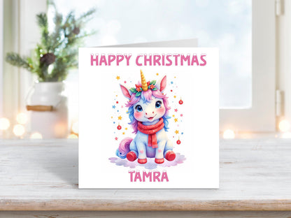 Personalised Christmas card featuring a pastel unicorn with a red scarf and holly headpiece, surrounded by festive baubles and stars, with “Happy Christmas Tamra” in pink text.