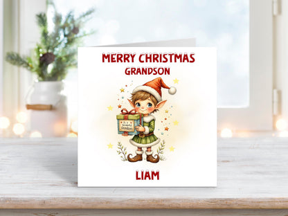 Christmas card for grandson featuring a festive elf in a green outfit and red hat holding a wrapped gift with “To A Wonderful Grandson” on the tag.
