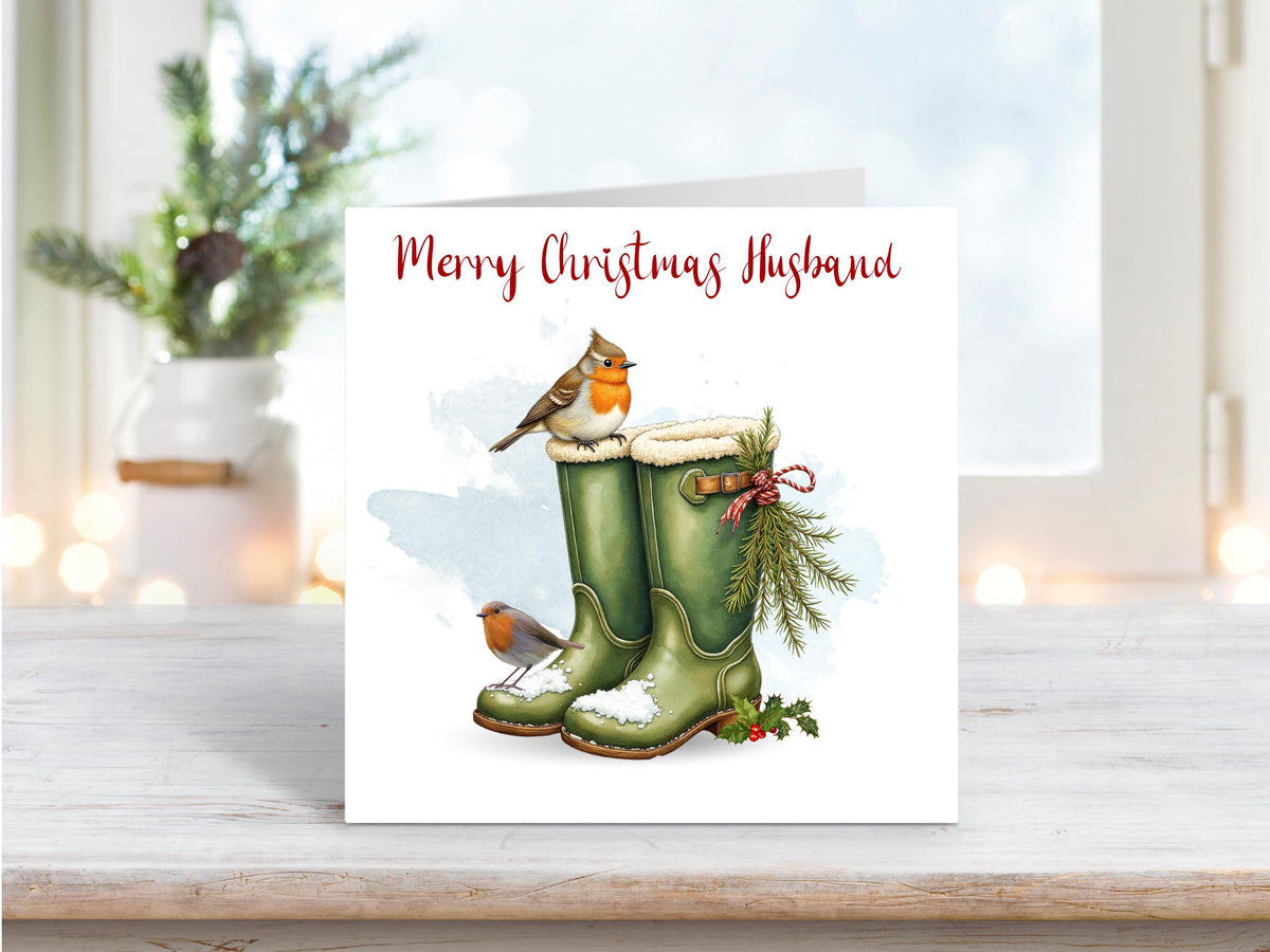 Christmas card for husband featuring green wellington boots in snow with two robins and festive foliage tied with red string.