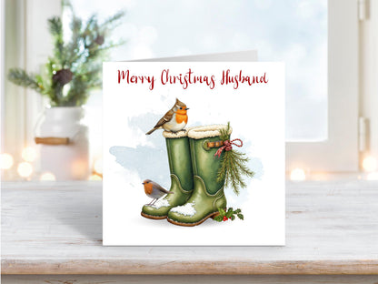 Christmas card for husband featuring green wellington boots in snow with two robins and festive foliage tied with red string.
