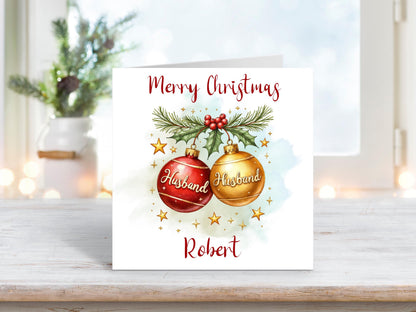 Christmas card for husband with two red and gold baubles labelled “Husband,” decorated with pine, holly, and stars, personalised with name underneath.