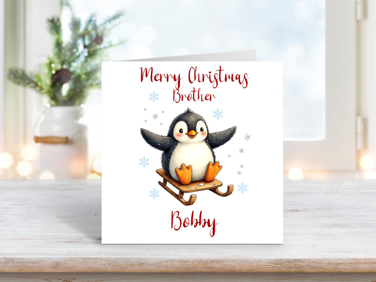 Christmas card with an adorable penguin riding a wooden sled, surrounded by snowflakes, with “Merry Christmas Brother” and personalised name “Bobby”.
