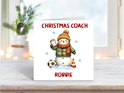 Festive Christmas card with a smiling snowman dressed as a football coach, holding a clipboard beside a football and training cone, with “Christmas Coach” and personalised name “Robbie”.