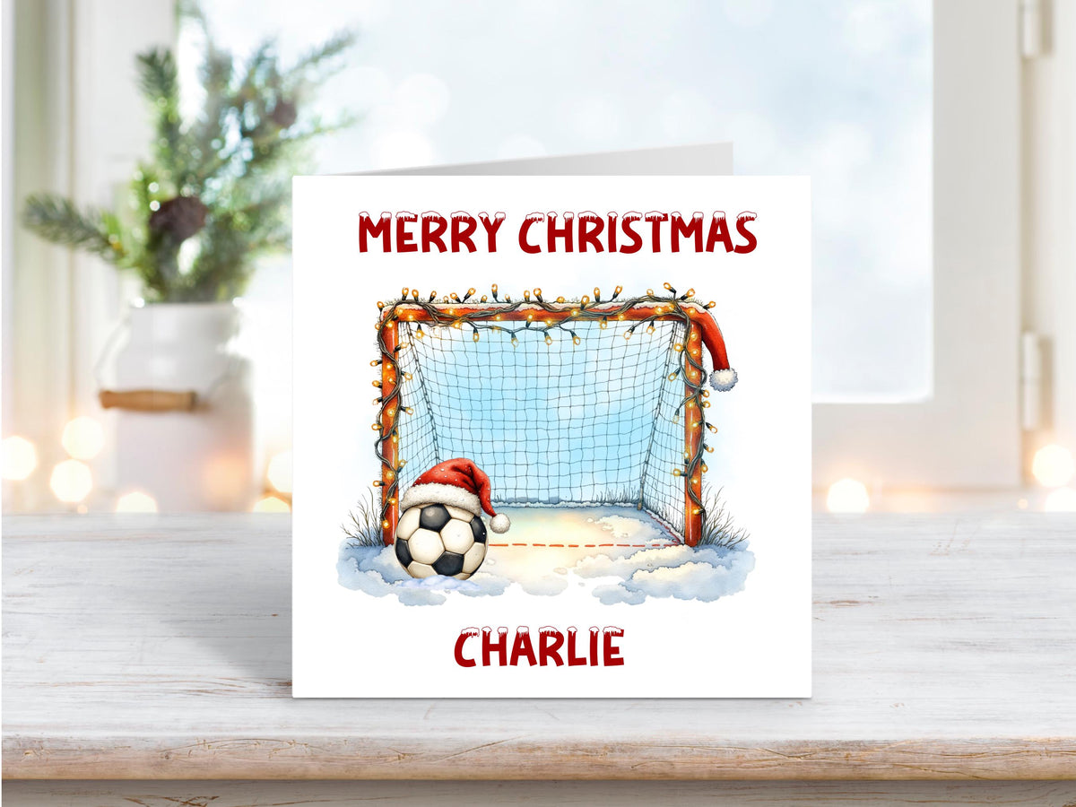 Christmas card showing a snowy football goal decorated with fairy lights and a Santa hat on a football, personalised with the name Charlie.