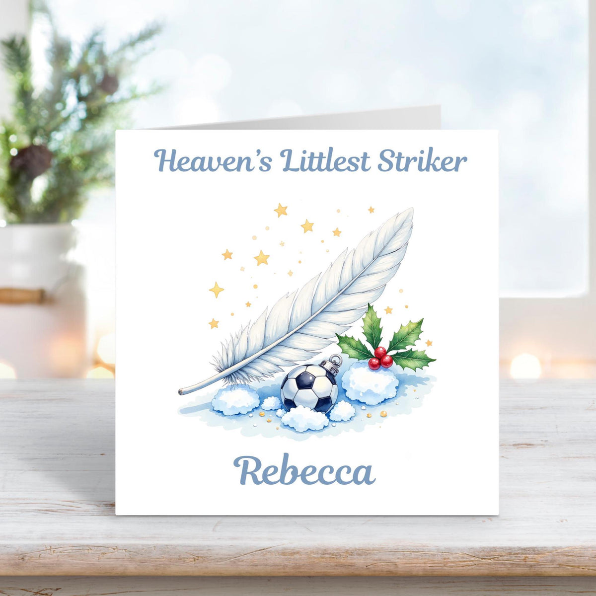 Memorial card featuring a soft white feather, holly leaves, and a small football resting in snow with golden stars, and the text “Heaven’s Littlest Striker Rebecca.”