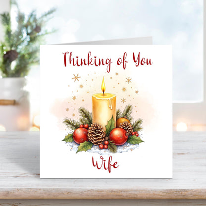 Christmas card for wife with “Thinking of You” message, featuring a glowing candle, pine cones, berries, and festive greenery.