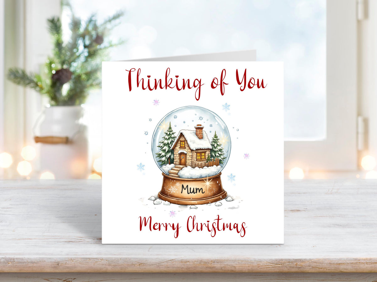 Christmas card featuring a snow globe with a cosy winter cottage and fir trees inside, with the words “Thinking of You Mum Merry Christmas” on a white background.
