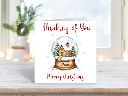 Christmas card featuring a snow globe with a cosy winter cottage and fir trees inside, with the words “Thinking of You Mum Merry Christmas” on a white background.