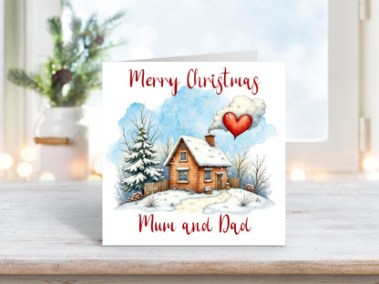 Christmas card featuring a snow-covered cottage with a red heart-shaped cloud in the sky, surrounded by frosted trees, with the words “Merry Christmas Mum and Dad.”