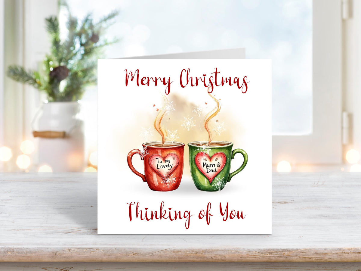 Christmas card featuring two steaming mugs, one red and one green, with heart labels reading “To My Lovely” and “Mum & Dad,” surrounded by snowflakes and the words “Merry Christmas Thinking of You.”