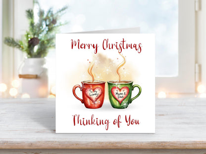 Christmas card featuring two steaming mugs, one red and one green, with heart labels reading “To My Lovely” and “Mum & Dad,” surrounded by snowflakes and the words “Merry Christmas Thinking of You.”