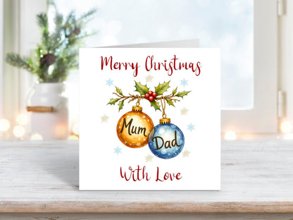 Christmas card featuring two hanging baubles labelled “Mum” and “Dad,” decorated with holly, berries, and snowflakes on a white background, with the words “Merry Christmas With Love.”