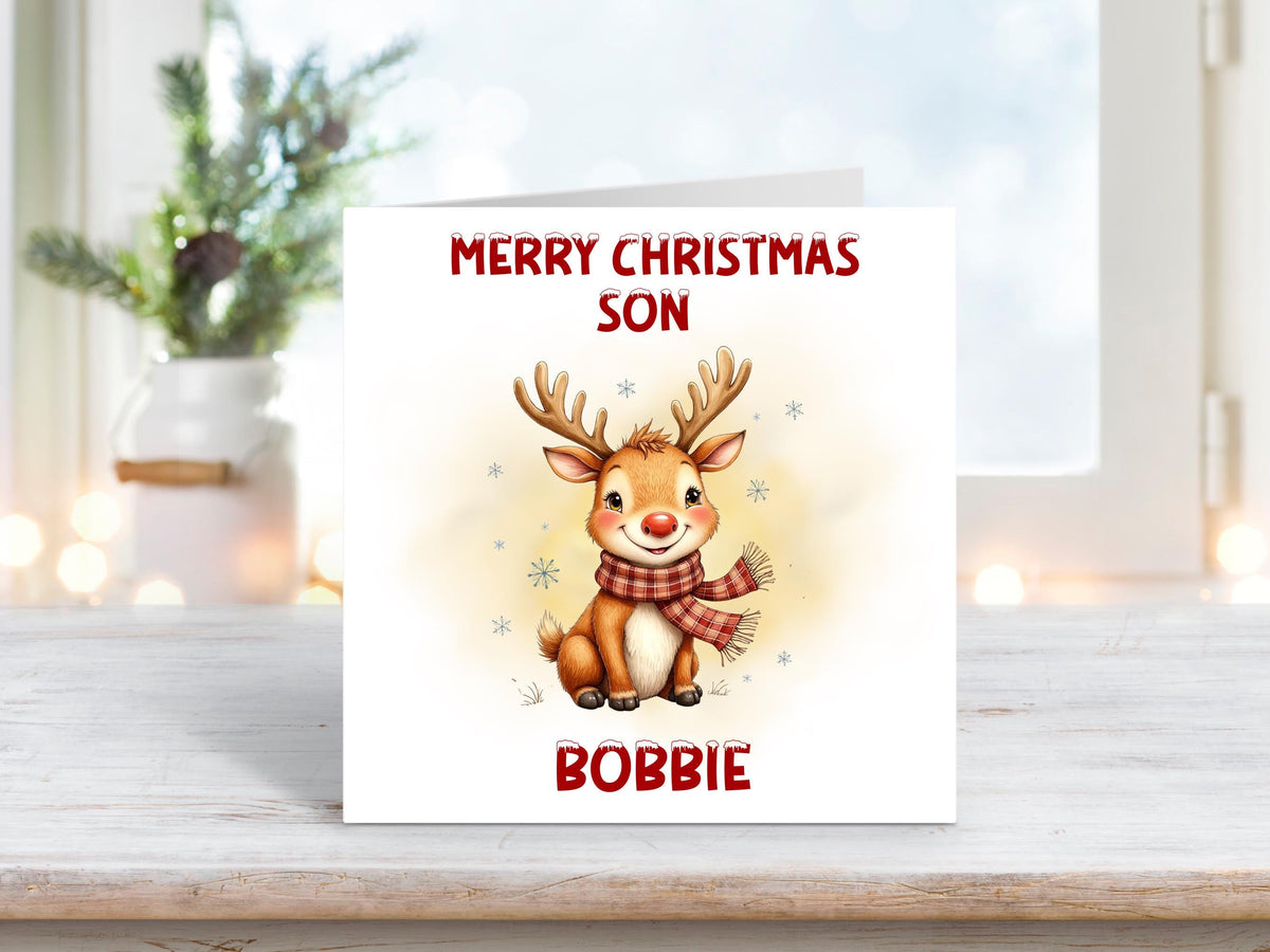 Christmas card featuring a smiling reindeer wearing a red scarf with snowflakes around it, and the words “Merry Christmas Son” and personalised name “Bobbie” on a white background.