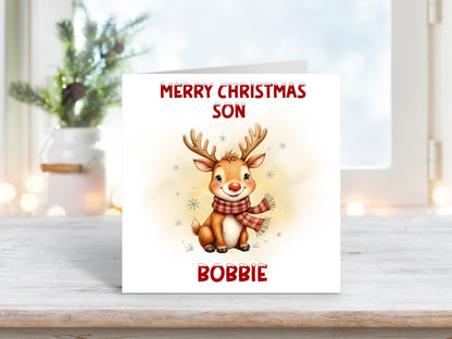 Christmas card featuring a smiling reindeer wearing a red scarf with snowflakes around it, and the words “Merry Christmas Son” and personalised name “Bobbie” on a white background.