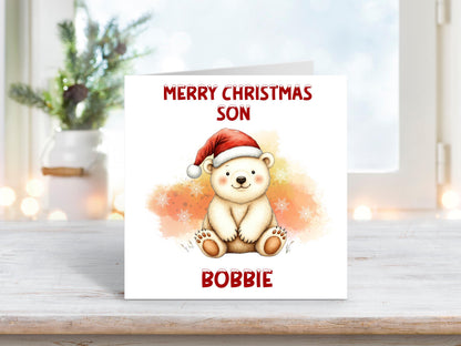 Christmas card featuring a smiling polar bear wearing a red Santa hat against a soft orange and yellow snowy background, with the words “Merry Christmas Son” and personalised name “Bobbie.”