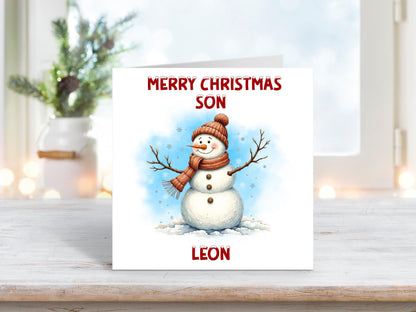 Christmas card featuring a happy snowman wearing a red hat and scarf with stick arms and snowflakes around, set against a blue snowy background, with the words “Merry Christmas Son” and personalised name “Leon.”