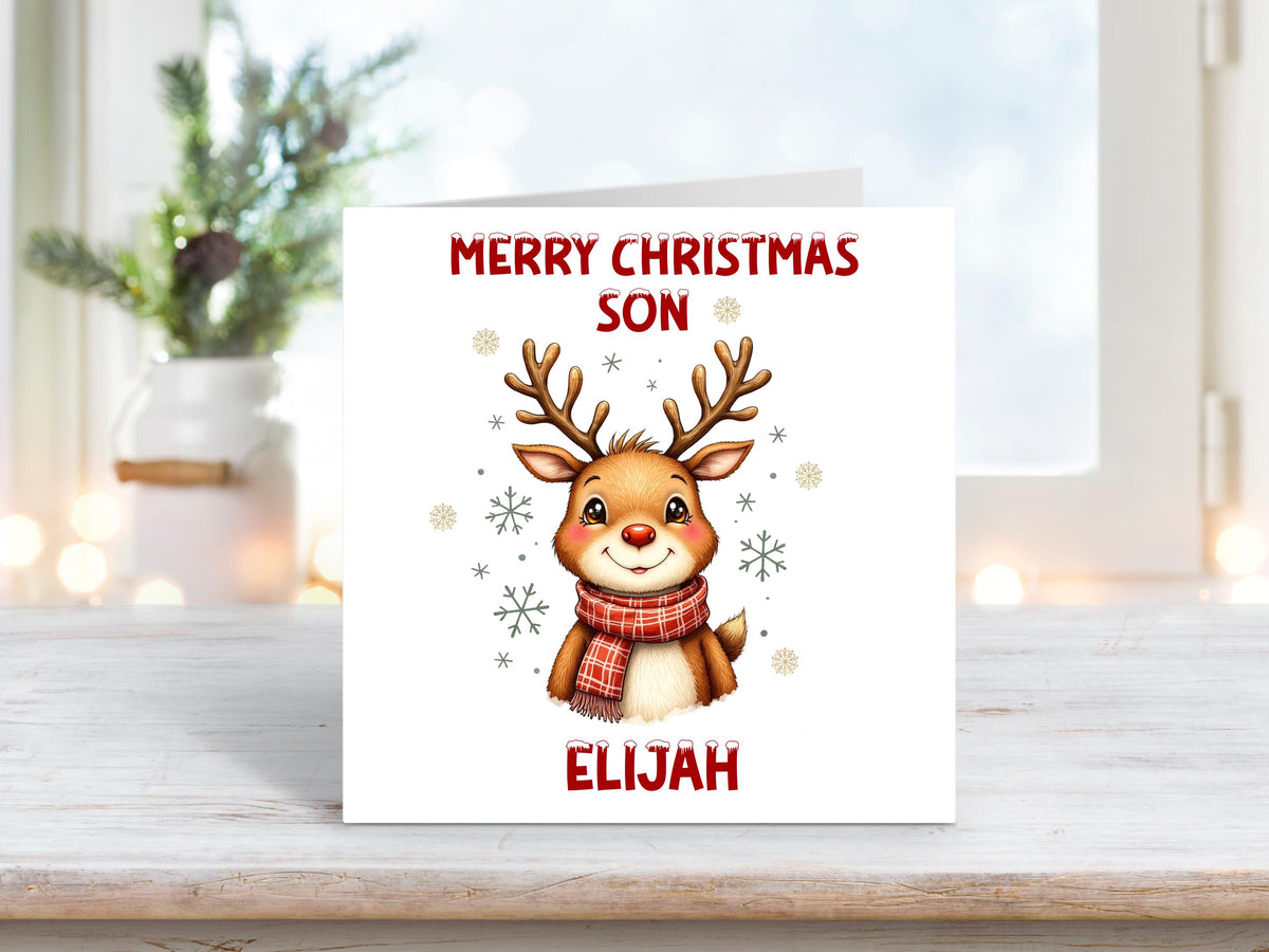 Christmas card featuring a smiling reindeer wearing a red scarf surrounded by snowflakes on a white background, with the words “Merry Christmas Son” and personalised name “Elijah.”