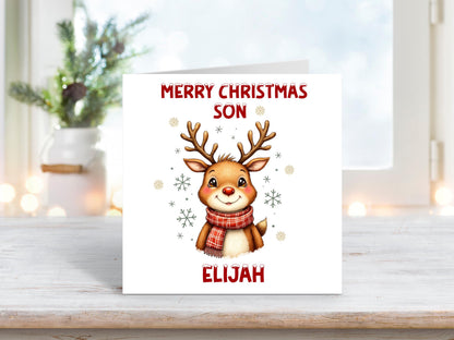 Christmas card featuring a smiling reindeer wearing a red scarf surrounded by snowflakes on a white background, with the words “Merry Christmas Son” and personalised name “Elijah.”