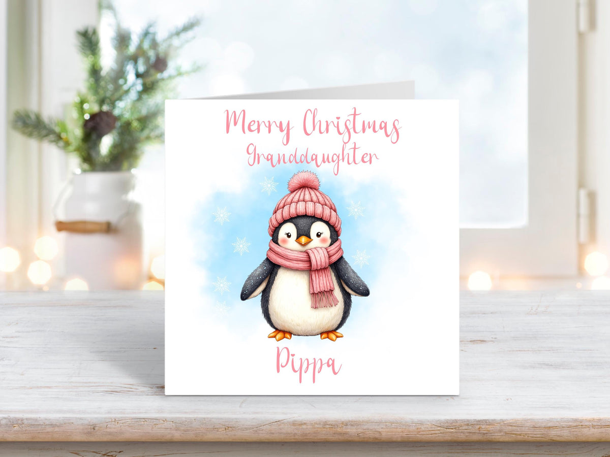 Personalised granddaughter Christmas card featuring an adorable penguin in a pink hat and scarf on a white background, surrounded by snowflakes.
