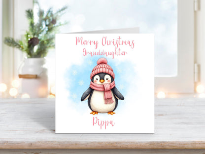 Personalised granddaughter Christmas card featuring an adorable penguin in a pink hat and scarf on a white background, surrounded by snowflakes.