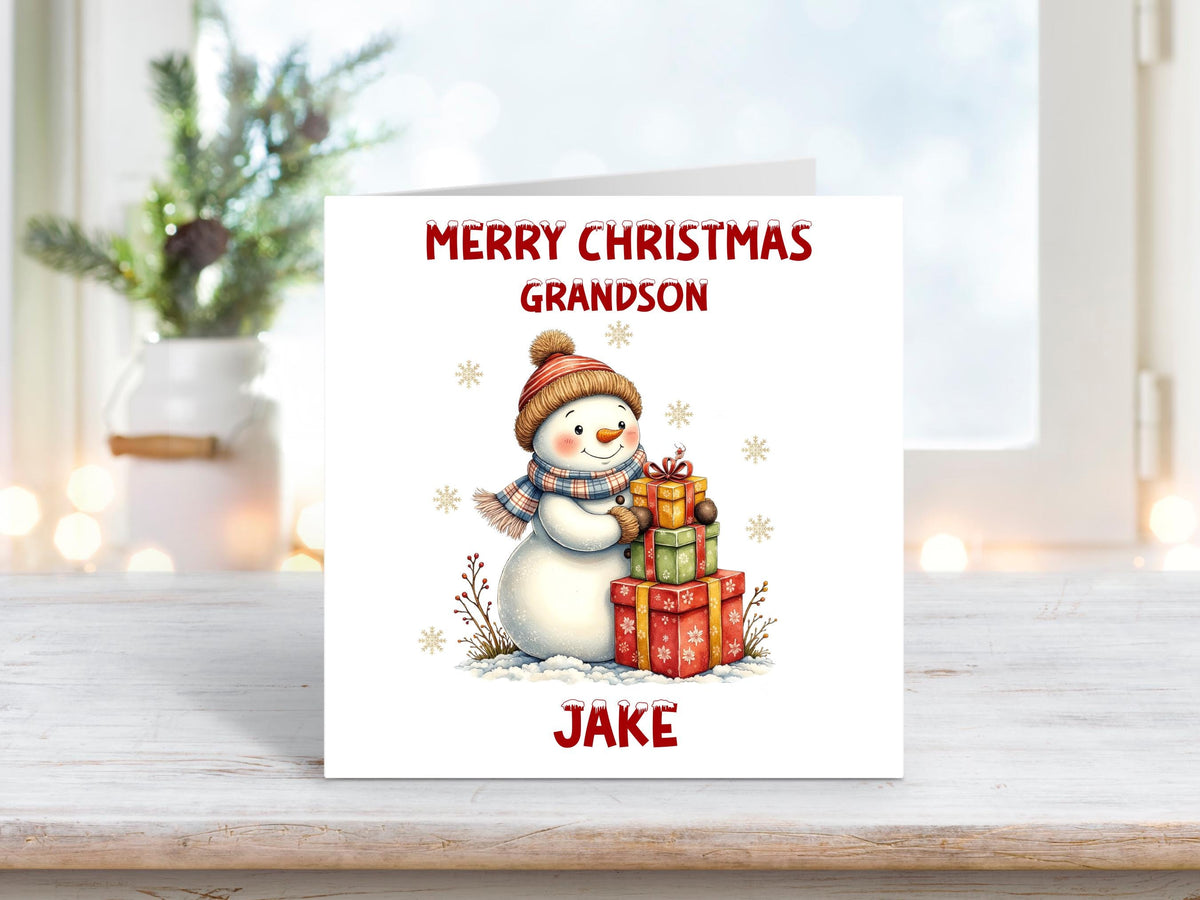 Christmas card for a grandson featuring a cheerful snowman wearing a hat and scarf, standing with a stack of wrapped gifts and personalised with a name.