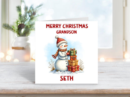 Personalised Christmas card for a grandson featuring a smiling snowman in a red hat and scarf, holding a stack of wrapped presents with festive greenery in the snow.