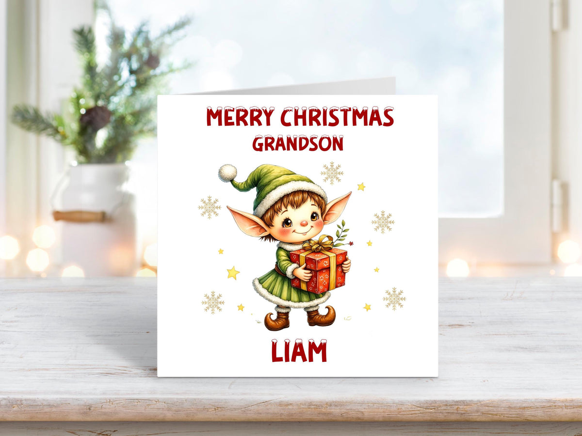 Personalised Christmas card for a grandson featuring a sweet elf in a green outfit and Santa hat, holding a red gift with golden ribbon and surrounded by snowflakes.