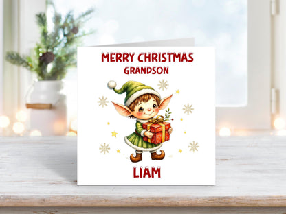 Personalised Christmas card for a grandson featuring a sweet elf in a green outfit and Santa hat, holding a red gift with golden ribbon and surrounded by snowflakes.