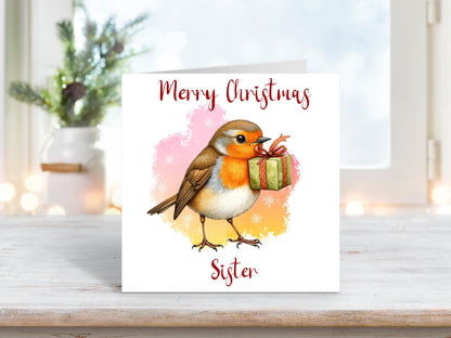 Christmas card for sister featuring a robin holding a green present in its beak, with a pink and yellow snowy background.