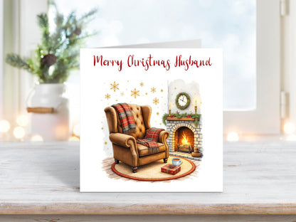 Christmas card for husband featuring a cosy armchair by a lit fireplace, with wrapped gifts, a mug, and festive decorations.