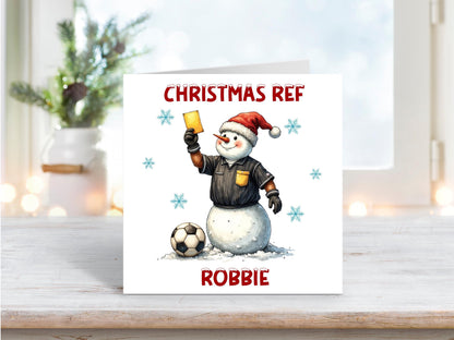Christmas card featuring a cheerful snowman referee wearing a Santa hat, holding a yellow card beside a football, with “Christmas Ref” and the personalised name Robbie.