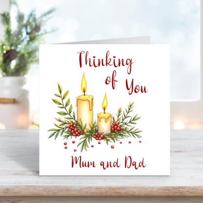 Christmas card featuring two glowing candles surrounded by holly, berries, and leaves, with the words “Thinking of You Mum and Dad” on a white background.