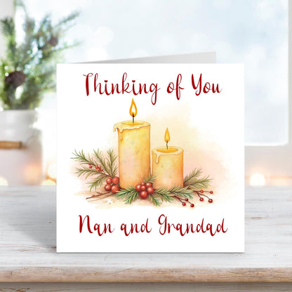 Christmas card featuring two glowing candles surrounded by holly, berries, and pine sprigs, with the words “Thinking of You Nan and Grandad” on a white background.
