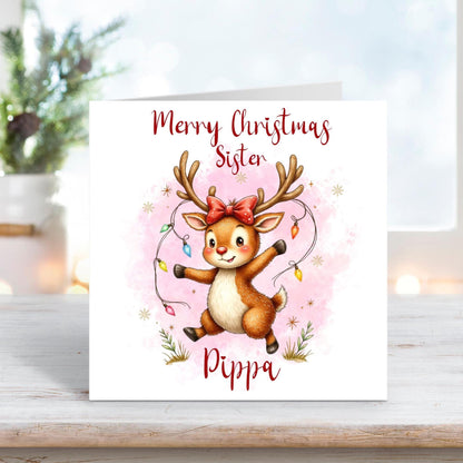 Christmas card for sister featuring a cute reindeer with a red bow and fairy lights wrapped around its antlers on a pink snowy background.